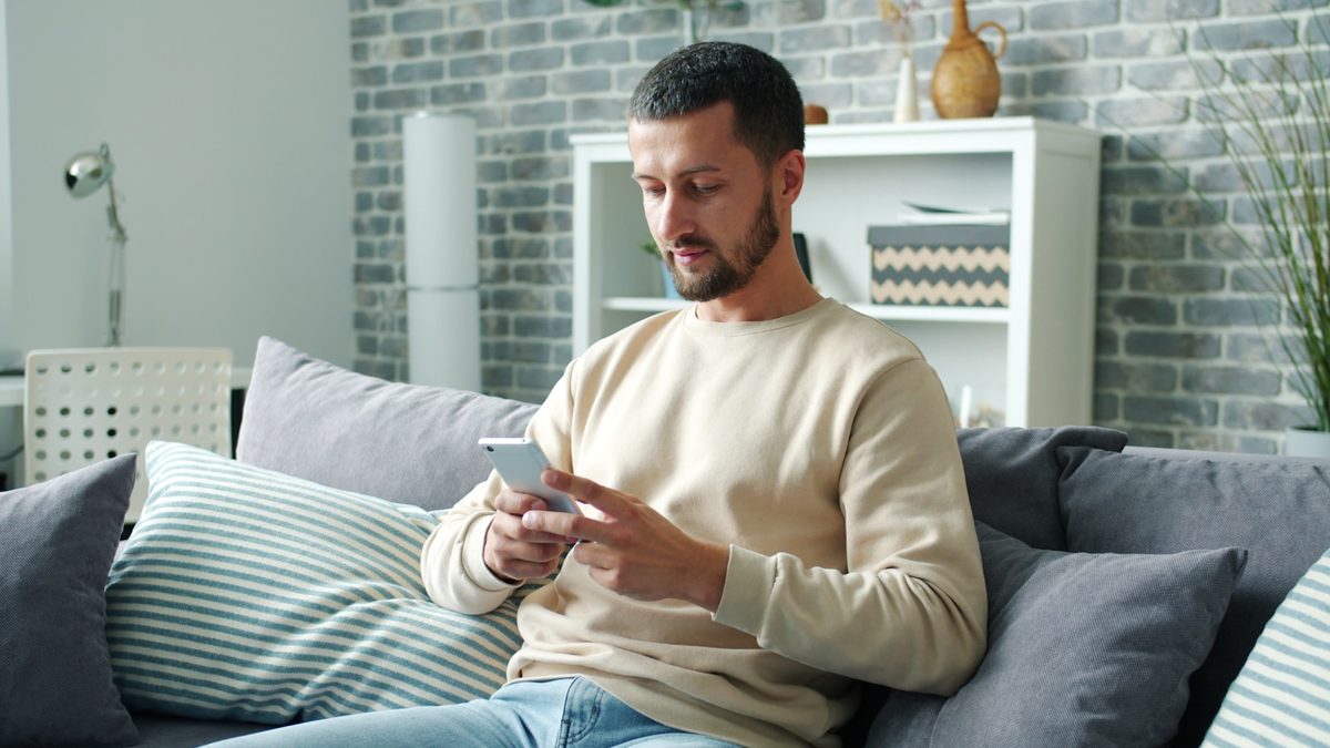 Mann sjekker opplysninger på mobilen hjemme i sofaen